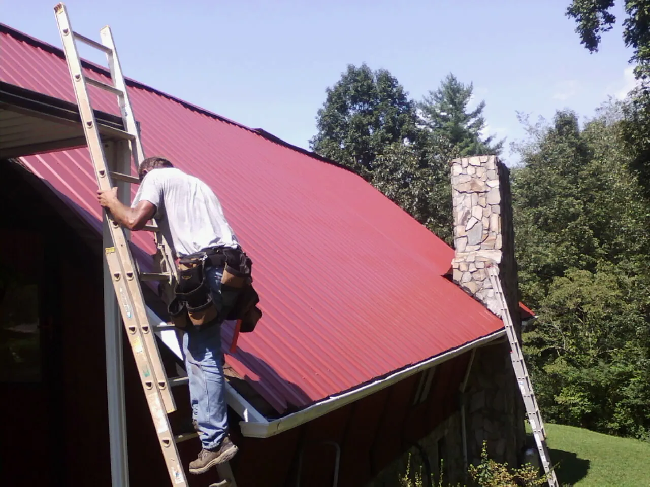 Roofer on a ladder beside a red metal roof and stone chimney — Soffit & Fascia Repair by Roofing Brothers in Pinole